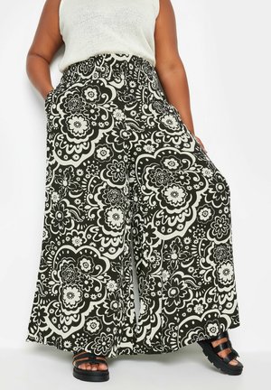 Person i plus-size iført sorte og hvide blomstermønstrede bukser med vide ben, hvid ærmeløs top og sorte sandaler, stående med hænderne i lommerne.