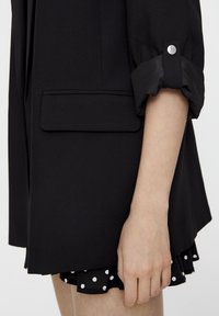 Black blazer with a smooth texture, featuring a front pocket, rolled cuffs with snap buttons, over a black polka-dotted skirt.