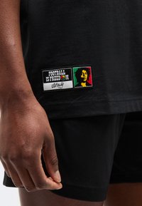 Κοντινό πλάνο του χεριού ενός ατόμου και ενός μαύρου πουκαμίσου που δείχνει ένα έμβλημα με το κείμενο «Το ποδόσφαιρο είναι ελευθερία», μια πολύχρωμη μπάλα ποδοσφαίρου και ένα στυλιζαρισμένο πρόσωπο σε κόκκινο, πράσινο και κίτρινο.