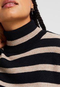 Pletený pruhovaný rolák v námořnické modři a béžové barvě, který má žebrovaný límec a texturovanou látku. Pohled zblízka na výstřih.