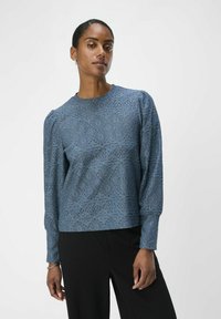 Haut en dentelle bleu à manches longues, col rond, motif texturé et coupe décontractée, associé à un pantalon noir.