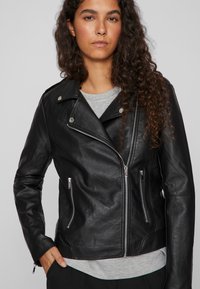 Black leather biker jacket with silver hardware, asymmetrical zipper, and two front zip pockets. Features a lightweight and slightly textured finish.