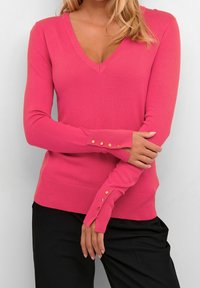 Różowy sweter z dekoltem w serek o dopasowanym kroju i długich rękawach. Zdobiony guzikami na mankietach oraz prążkowanym brzegiem dla dodatkowej tekstury.