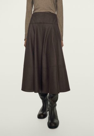 Femme portant une jupe midi taille haute marron foncé associée à des bottes noires montant aux genoux et un haut à manches longues couleur taupe.