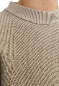 Pull en tricot de couleur taupe clair, présentant une texture côtelée, un col montant et une transition sans couture au niveau de l'épaule.