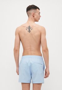 Lys blå badebukser med strukturert finish, elastisk midje og en baklomme. Modellen har en tatovering på den øvre delen av ryggen.