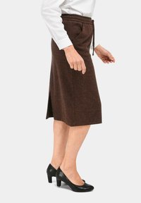 Jupe brune en chevrons avec une taille à nouer, de longueur mi-mollet, avec une fente sur le côté. Portée avec des chaussures à talons hauts noires.