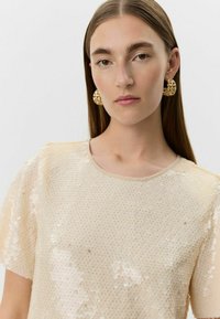 Kvinde med glat brunt hår iført en beige paillet-top og store, teksturerede guldringøreringe mod en ensfarvet hvid baggrund.