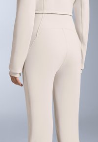 Beżowy dopasowany kombinezon z długimi rękawami, gładką fakturą oraz widocznymi szwami z tyłu i po bokach, podkreślającymi sylwetkę.