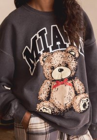 Harmaa collegepaita, jossa on nalletiketti punaisella solmulla ja "MIAMI" -teksti paksulla, valkoisella kirjaimella. Pehmeä kangas ja rento malli.