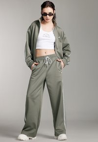 Olivgrüner Trainingsanzug mit weißen Streifen. Die Jacke hat einen Reißverschluss auf der Vorderseite und einen hohen Kragen, während die Hose einen elastischen Bund und Taschen hat.