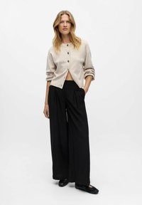 Donna con lunghi capelli biondi, indossa un cardigan beige con bottoni, pantaloni neri a gamba larga e scarpe nere, in piedi davanti a uno sfondo bianco semplice.