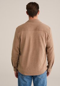 Ruda tekstūruota marškiniai su apykakle, ilgomis rankovėmis ir mygtuko akcentu ant rankogalių, derinami su šviesiai mėlynomis nusidėvėjusiomis džinsomis.