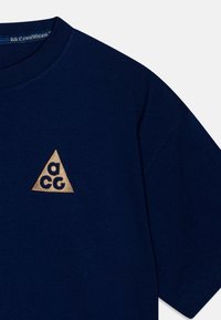 Jūras spēku kokvilnas T-krekls ar zelta trīsstūra logotipu un "aCG" tekstu. Apkaklīte ir ribotas formas, un audumam ir gluda tekstūra.