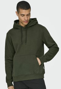 Felpa con cappuccio in verde oliva scuro, realizzata in tessuto morbido, con tasca a marsupio frontale, cordini regolabili e polsi a costine.