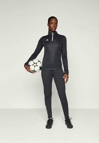 Svart atletisk topp med kvart glidelås og Adidas-logo, kombinert med svarte tettsittende bukser med hvite sidedetaljer. Holder en fotball.