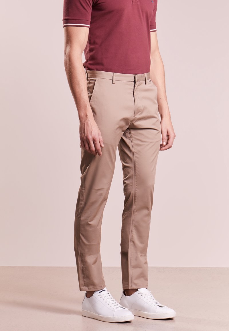 Chino beige con un design slim fit, caratterizzati da una texture liscia e tasche standard. Indossati con una polo bordeaux e sneakers bianche.