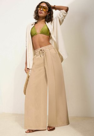 Kvinde iført olivengrøn bikini top, beige bukser med vide ben, offwhite åben skjorte, gultintede solbriller, der holder en vævet taske, poserer mod en ensfarvet baggrund.