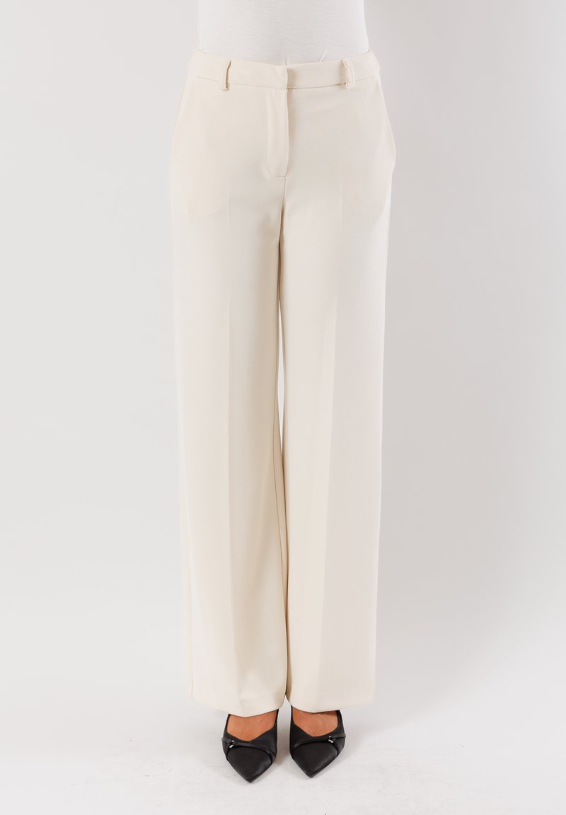 Pantaloni a gamba larga in tessuto crema con una consistenza liscia, caratterizzati da una vita media, pieghe e tasche laterali. Abbinati a tacchi neri a punta.