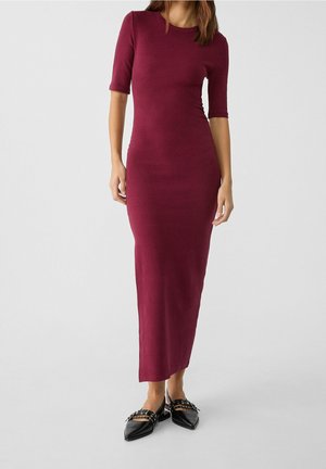 Kobieta ubrana w dopasowaną, ciemnoczerwoną sukienkę midi z rękawami do łokcia oraz czarne, zaokrąglone baleriny na pasku wokół kostki z ozdobnymi klamrami.