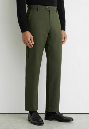 Herrenhose in Olivgrün, gerade geschnitten, kombiniert mit schwarzen Loafers und einem schwarzen Langarmshirt, dargestellt von der Taille bis zu den Füßen.