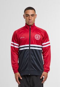 Rød og marineblå lynlås sportsjakke med hvide striber, ribbede manchetter og broderet logo på brystet. Fremstillet af glat syntetisk materiale.