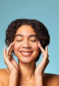 Nahaufnahme einer Person mit lockigen schwarzen Haaren und Sommersprossen, die lächelt und sanft ihr Gesicht berührt. Weicher Hautton vor einem hellblauen Hintergrund.