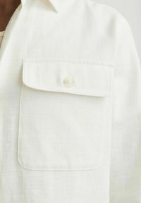 Camisa de lino blanca con un bolsillo de solapa y cierre de botón. La tela tiene una apariencia texturizada con una calidad ligera.