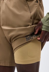 Σκούρος σε επιδερμίδα άτομο σηκώνει μπεζ σορτς για να αποκαλυφθεί στενό μπεζ εσώρουχο με μικρή τσέπη, φοράει πράσινο μακρυμάνικο μπλουζάκι.