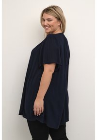 Donna sorridente con capelli biondi, che indossa una maglietta blu navy ampia e pantaloni neri, in piedi contro una parete chiara e semplice.