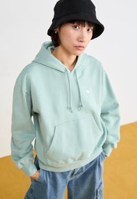 Sweat à capuche vert menthe avec une poche avant et des cordons réglables, porté avec un pantalon en denim ample. Un chapeau seau noir ajoute du contraste.
