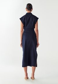 Abito midi blu navy con maniche corte strutturate, vita alta e spacchi laterali. Texture liscia e vestibilità sartoriale, abbinato a sandali con tacco beige.