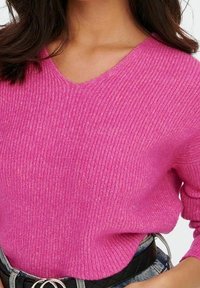 V-Ausschnitt, gerippter Strickpullover in leuchtendem Pink mit langen Ärmeln. Wird über einem schwarzen Gürtel mit rundem silbernem Schnallenverschluss getragen.