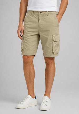 Homme portant un short cargo beige avec des poches latérales, associé à des baskets blanches et un t-shirt blanc, debout devant un fond uni.