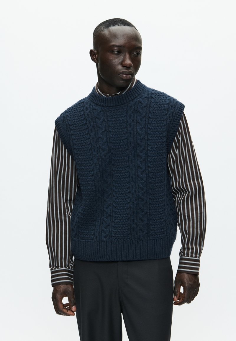 Sötétkék kötött mellény magas nyakkal és texturált zsinórmintákkal, egy barna és fehér csíkos hosszú ujjas pólóra rétegezve.