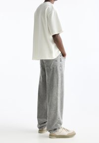 Personne portant un t-shirt blanc surdimensionné, un jean gris ample et des baskets beiges, debout les mains dans les poches devant un fond blanc.