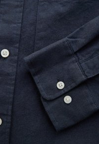 Námornícky modrá košeľa s jemnou textúrou, s detailom gombíkov na manžete a nápleci. Gombíky sú guľaté a biele s dvoma malými dierkami.