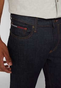 Gros plan sur une personne portant un jean bleu foncé avec des coutures orange et un haut beige clair, montrant la main gauche près de la hanche.