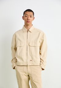Beige zip-up jakke med to frontlommer, spiss krage og lange ermer, laget av glatt stoff; brukt sammen med matchende beige bukser.