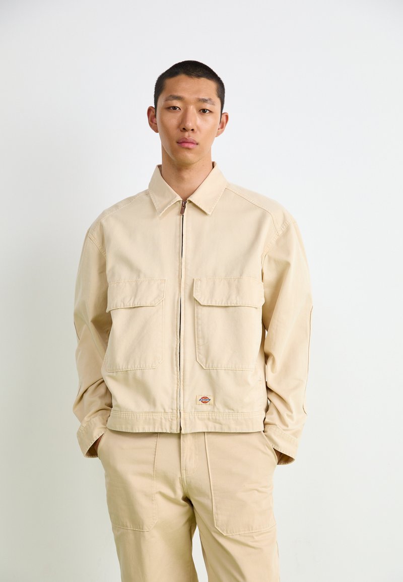 Beige zip-up jakke med to frontlommer, spiss krage og lange ermer, laget av glatt stoff; brukt sammen med matchende beige bukser.