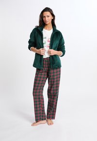 Grüne samtige Zip-Jacke über einem weißen T-Shirt mit Grafiken, kombiniert mit rot und grün karierten Schlafanzughosen. Modell barfuß vor einem schlichten Hintergrund stehend.