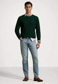 Suéter de malha em verde-escuro sobre uma camisa de colarinho rosa-claro, combinado com jeans azul-claro e sapatos de couro castanhos. Design simples e clássico.