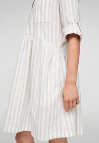 Robe rayée en beige clair et blanc. Elle présente un col à boutons, des manches courtes retroussées et une jupe froncée avec une coupe décontractée.