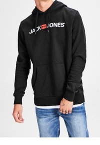 Man som bär en svart Jack & Jones hoodie med en framficka och slitna blå jeans framför en enfärgad bakgrund.