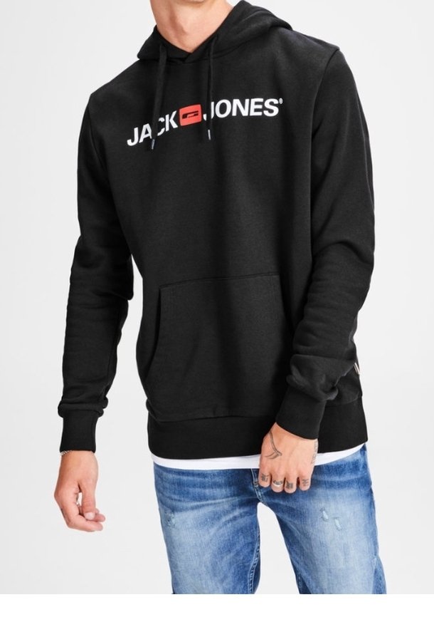 Man som bär en svart Jack & Jones hoodie med en framficka och slitna blå jeans framför en enfärgad bakgrund.