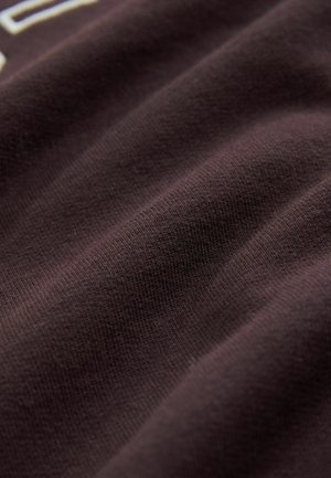 Tumši brūns audums ar gludu tekstūru, uz kura redzams smalks adījuma raksts un maigi krokas. Gaisma maigi atspoguļojas uz virsmas.