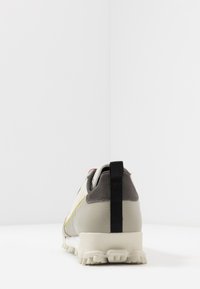 Sneaker med en ljusgrå ovandel, svarta accenter, textilmaterial och gula sömmar; har en sportig sula med multidirektionella klor.