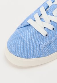 Sneaker in tela azzurra chiara con un motivo testurizzato, lacci bianchi e suola in gomma bianca con occhielli blu. Dettaglio ravvicinato della punta.