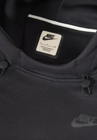 Crna dukserica s glatkom teksturom, s Nike oznakom, podešivim vezicama i bijelim naglaskom na zipu oko vrata.