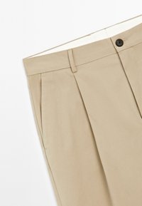 Beige trousers with pleats, made of smooth fabric, featuring a black button and a flat waistband, shown with a white background.
